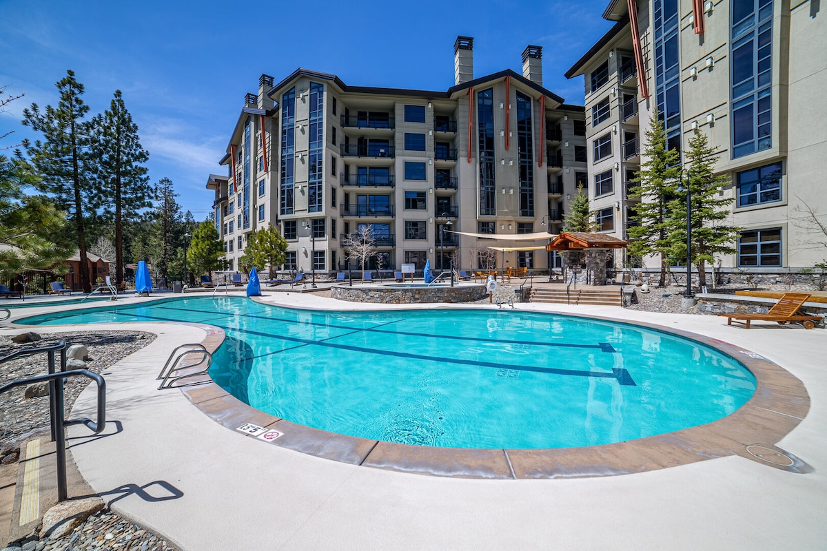 The outdoor heated pool is surrounded by lounge chairs and trees, offering a spacious area for relaxation. The modern multi-story building in the background features large windows and a welcoming entrance, creating a pleasant atmosphere for guests.