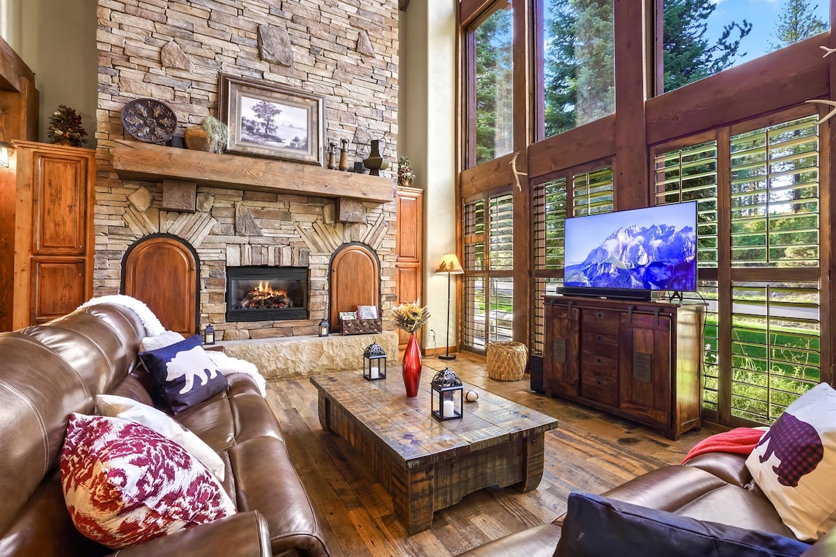 A spacious living area is highlighted by tall windows allowing natural light and views of the surrounding trees. A stone fireplace features prominently, complemented by a wooden coffee table and comfortable seating. Decorative pillows and lanterns add subtle warmth to the atmosphere.