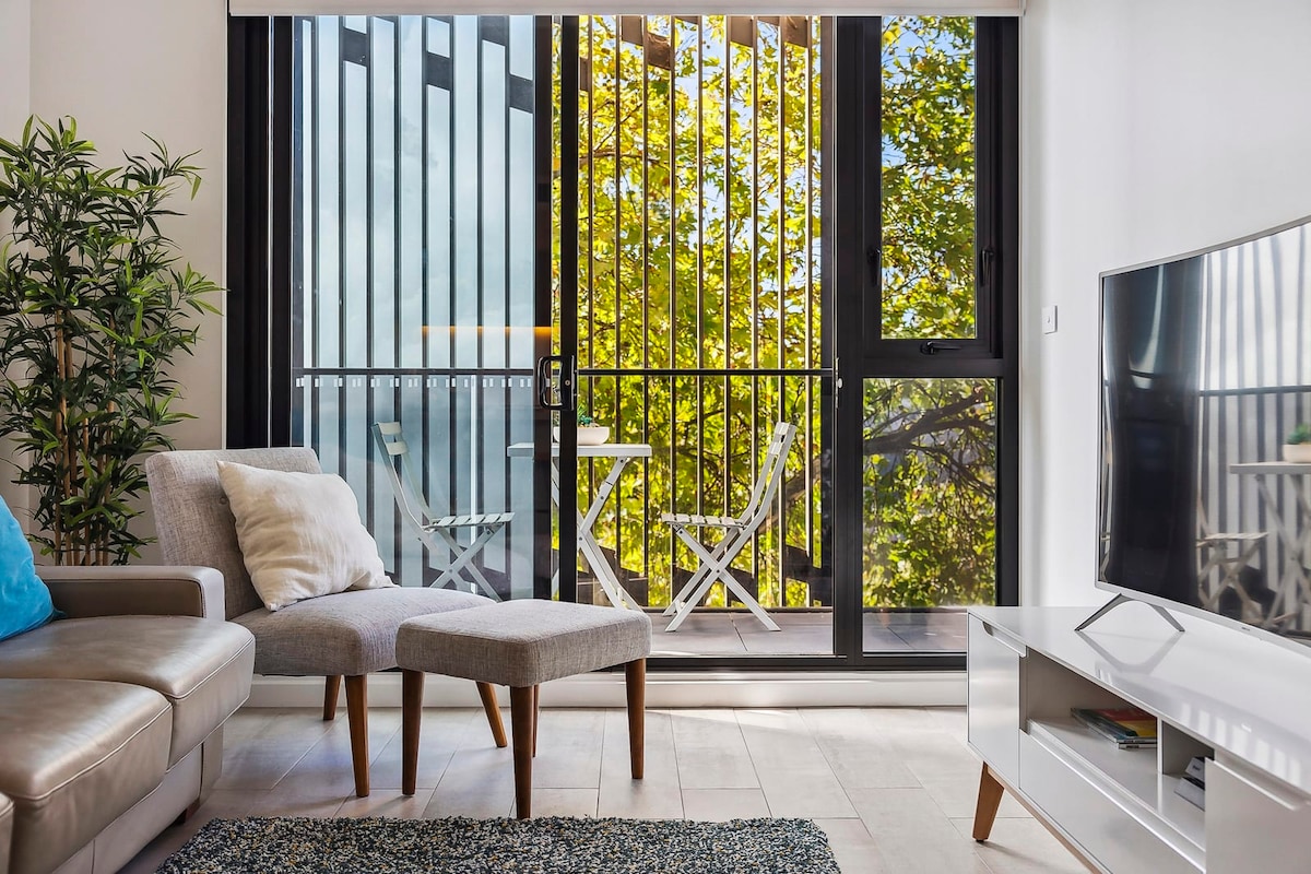 A modern living area features a comfortable sofa and ottoman, with a coffee table in front. Large glass doors open to a private balcony, where a small table and two chairs are visible among lush greenery. Natural light brightens the space.