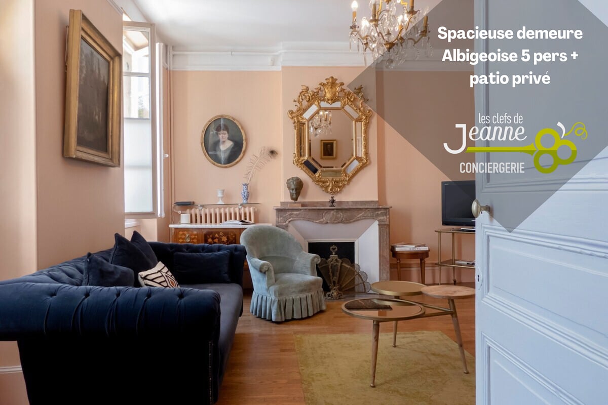 A spacious living area features elegant furnishings, including a blue couch and a vintage armchair. A decorative fireplace is complemented by a large mirror, adding a touch of sophistication. Natural light illuminates the room through tall windows, enhancing the welcoming ambiance.
