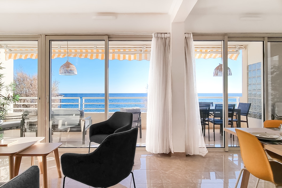 A spacious living area offers views of the sea through large glass doors. Outdoor dining space is visible with a set of black chairs and table on the terrace. Natural light fills the room, complemented by white curtains and modern furnishings.
