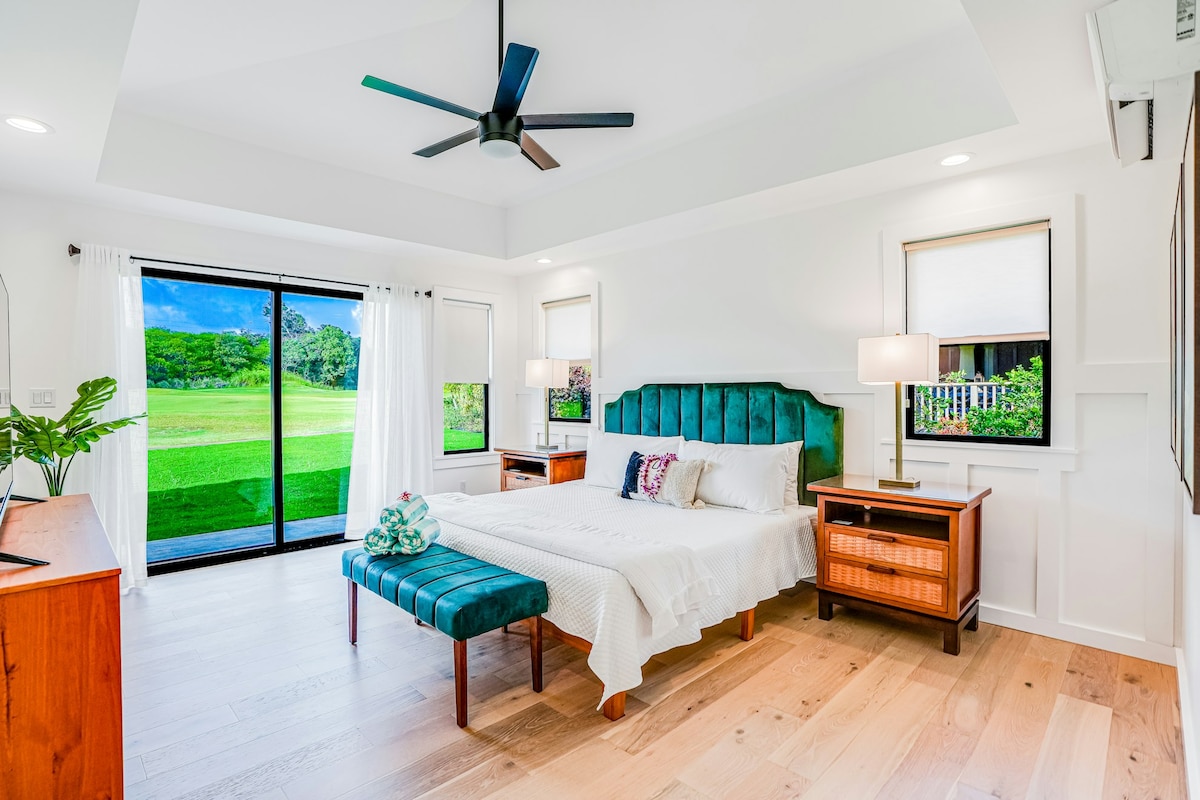 A spacious bedroom is illuminated by natural light, featuring a comfortable bed with a teal tufted headboard and white bedding. A wooden bench sits at the foot of the bed. Large glass doors open to views of green landscaping, and light-colored curtains frame the windows.