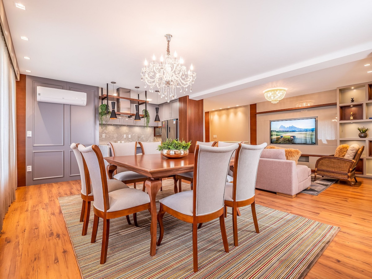 A spacious living and dining area features a large wooden dining table surrounded by six upholstered chairs. A crystal chandelier hangs above, providing soft lighting. The room includes a comfortable seating arrangement and a smart TV set against a textured wall, creating an inviting ambiance.