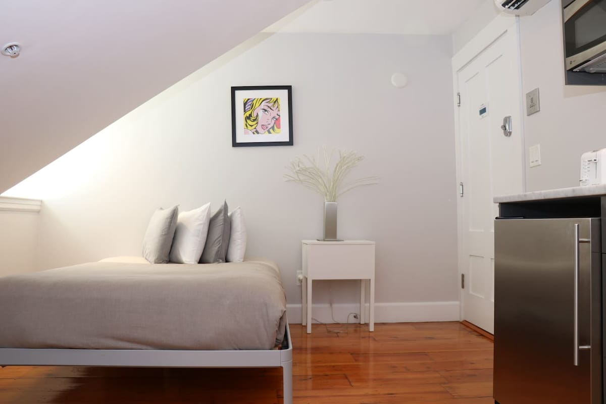 A cozy sleeping area features a spacious bed with soft grey linens and several pillows. A small nightstand holds a decorative vase with faux greenery. A framed artwork is displayed on the wall, and a kitchenette is visible in the corner, enhancing functionality.