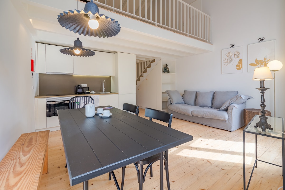 An open living area features a black dining table with four chairs and a soft gray sofa. Modern pendant lights hang above the table, illuminating the space. A staircase leads to an upper level, while light wood flooring complements the bright, neutral walls.