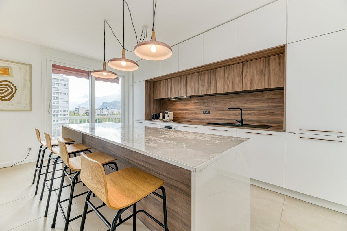 A modern kitchen features a spacious island with a light-colored countertop and wooden cabinetry. Four wooden stools are positioned along one side of the island. Three pendant lights hang above, illuminating the area, while a large window offers a view of the surrounding landscape.