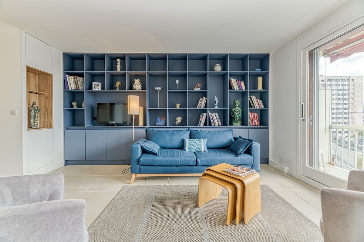 A modern living area is highlighted by a blue shelving unit filled with decor. A comfortable sofa sits in front of a compact television, while a stylish wooden coffee table is positioned in the center. Large glass doors provide access to a balcony, allowing for natural light.