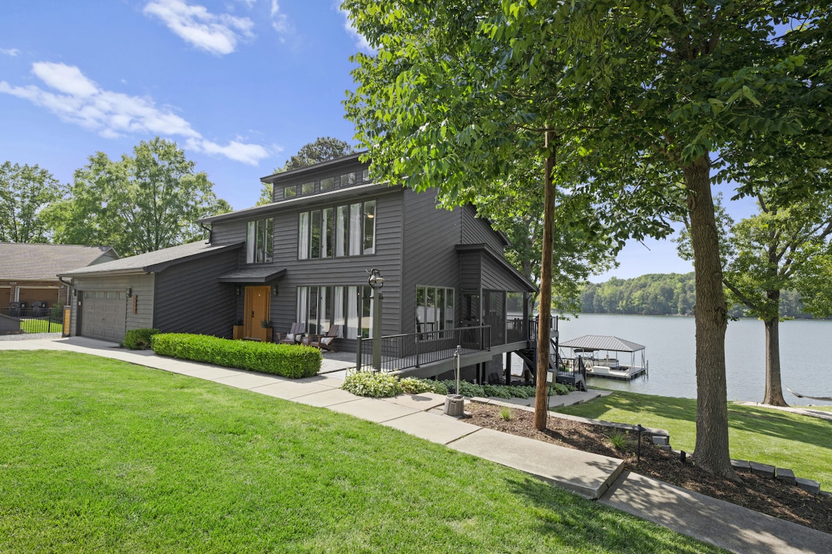 A modern two-story home with dark exterior is set against a backdrop of lush greenery and water. Large windows offer views of Lake Norman, while a covered deck extends from the side. A manicured lawn borders a pathway leading toward the lakefront.