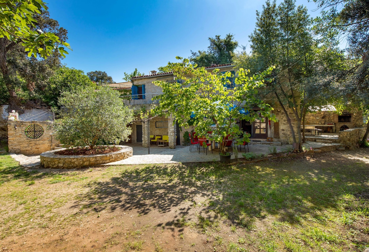 The stone holiday house is nestled within a lush garden, featuring mature trees and open grassy areas. A circular flower bed adds a pop of color, while outdoor seating is visible under a large tree. The facade of the house reveals multiple doors and windows, enhancing its welcoming appearance.