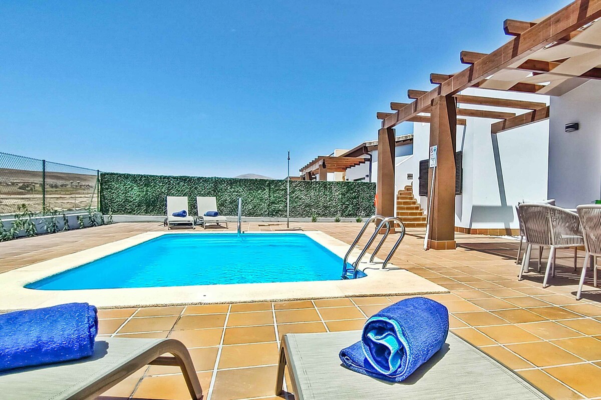An inviting outdoor pool is framed by a tiled terrace and surrounded by greenery. Two lounge chairs with blue towels are positioned nearby, enhancing relaxation options. A covered dining area is visible in the background, along with stairs leading to an upper terrace.