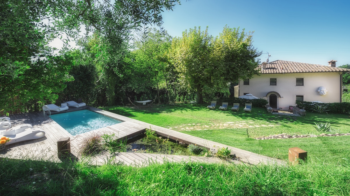A serene garden space features a rectangular swimming pool surrounded by a wooden deck. Comfortable sunbeds are positioned nearby, with grassy areas and trees providing shade. The charming house with a tiled roof and arched windows is seen in the background, completing the peaceful outdoor setting.