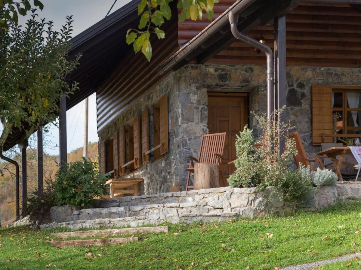 The exterior of the holiday home is shown, featuring a stone facade and wooden accents. A welcoming porch area is highlighted, with wooden chairs positioned beside lush greenery and a staircase leading down to the grassy yard.