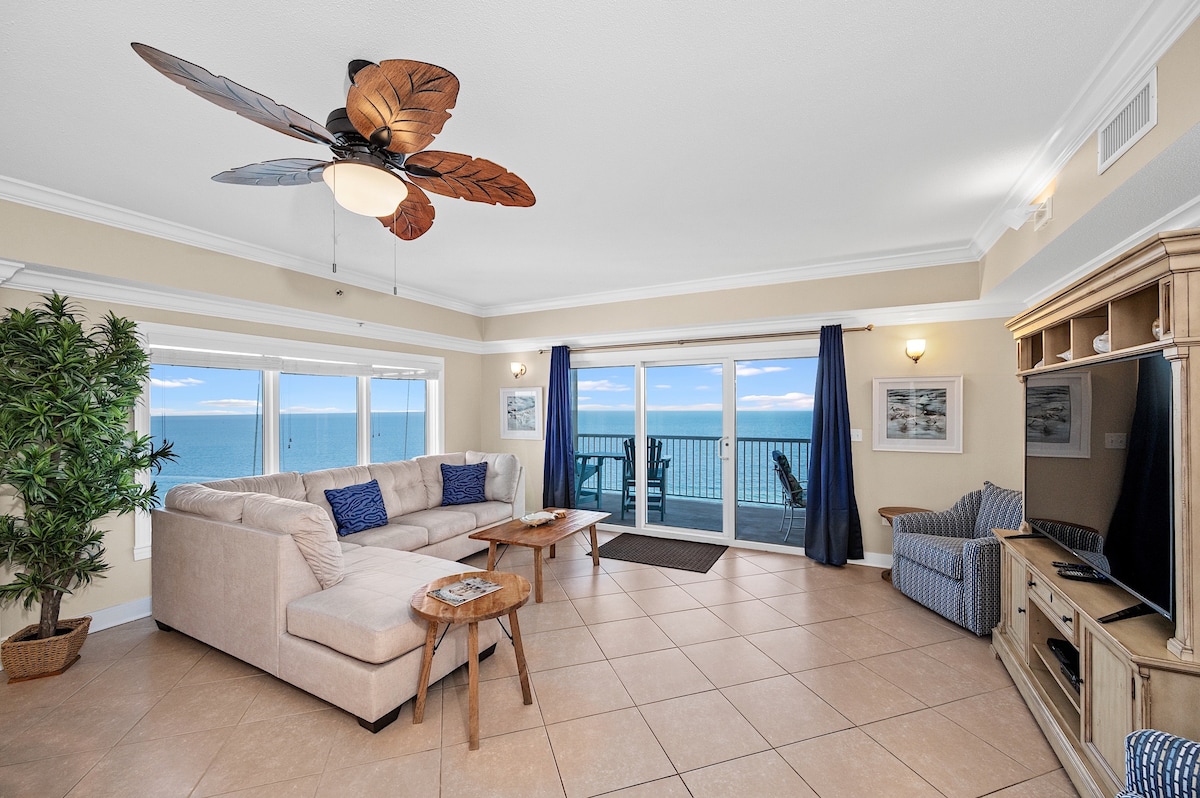 A spacious living area is seen featuring a sectional sofa and two wooden tables. Large windows allow natural light to fill the room and provide direct beachfront views. A ceiling fan with leaf-shaped blades adds a coastal touch, and the space is complemented by neutral-colored walls.