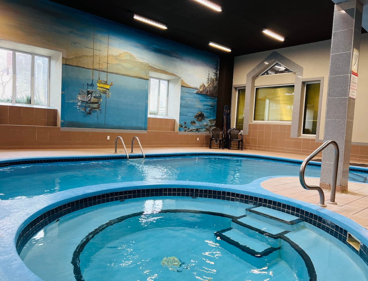 An indoor swimming pool with a hot tub is depicted, surrounded by a colorful mural of boats and a scenic landscape. Large windows allow natural light to illuminate the blue water, enhancing the tranquil atmosphere of the space.