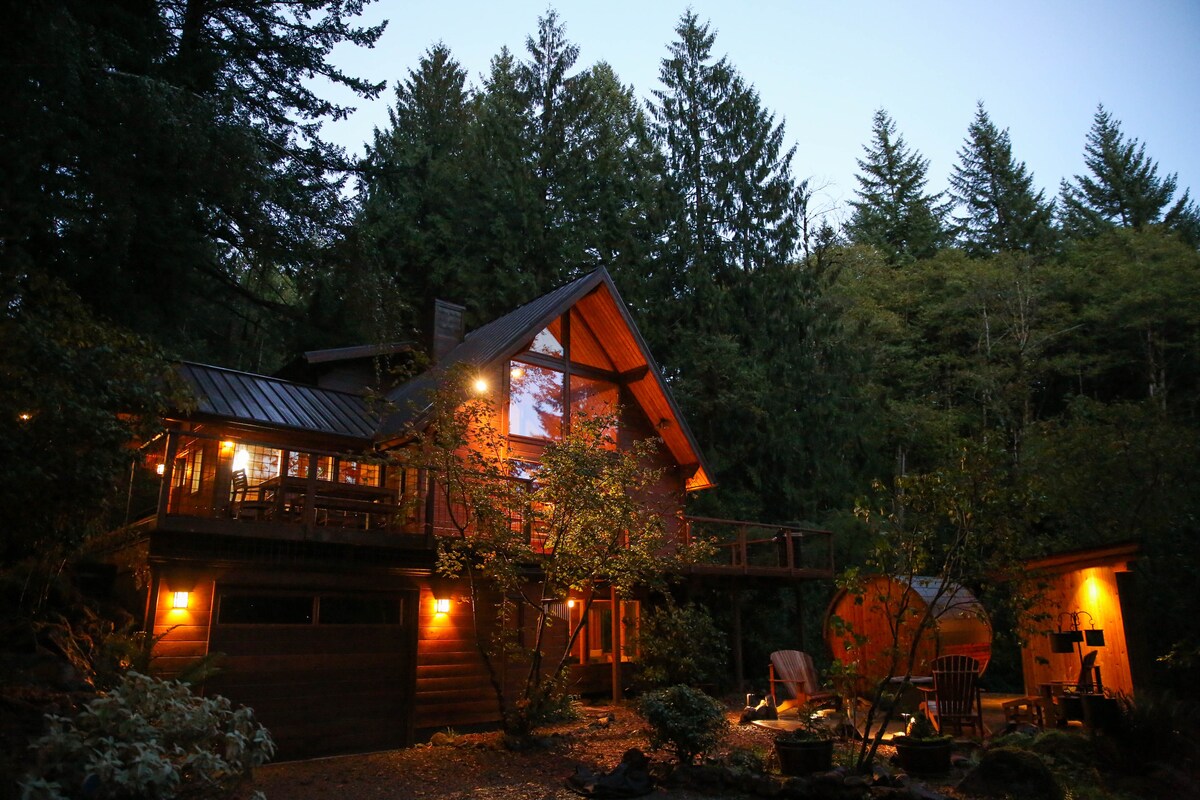 The A-frame cedar cabin is illuminated against the dusk sky, surrounded by towering evergreens. Soft lighting enhances the inviting exterior, featuring a spacious deck and outdoor seating areas. Natural elements blend with cozy features, creating a serene atmosphere in this forest retreat.