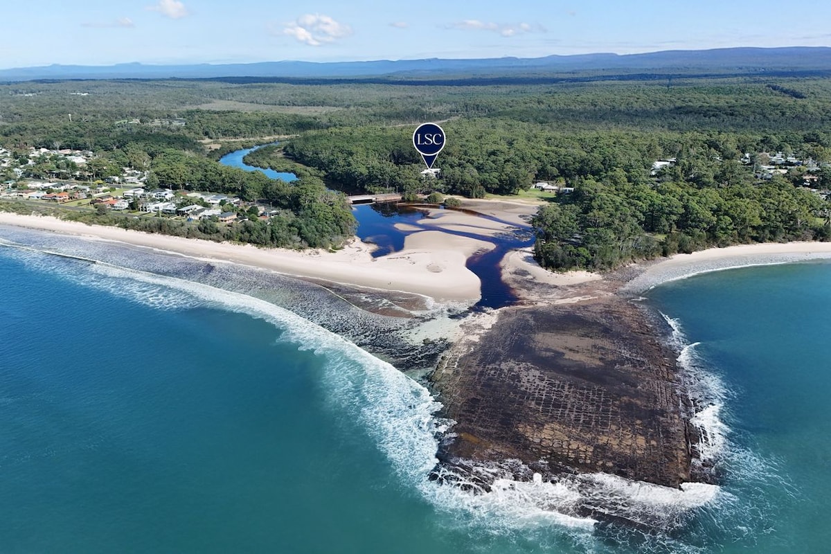 An aerial view reveals Moona Moona Creek meeting the coastline, with a sandy beach and rocky formations in the foreground. Lush green trees extend into the background, contrasting with the blue waters. The scene captures the natural beauty of the area, emphasizing serene outdoor settings.