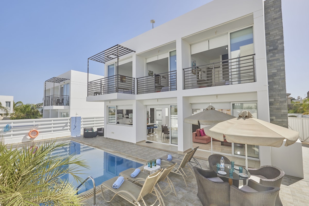The modern villa showcases a clear blue pool surrounded by comfortable sun loungers and shaded seating areas. Balconies off the upper-level rooms overlook the outdoor space, while large glass doors provide a seamless view of the interior living area.