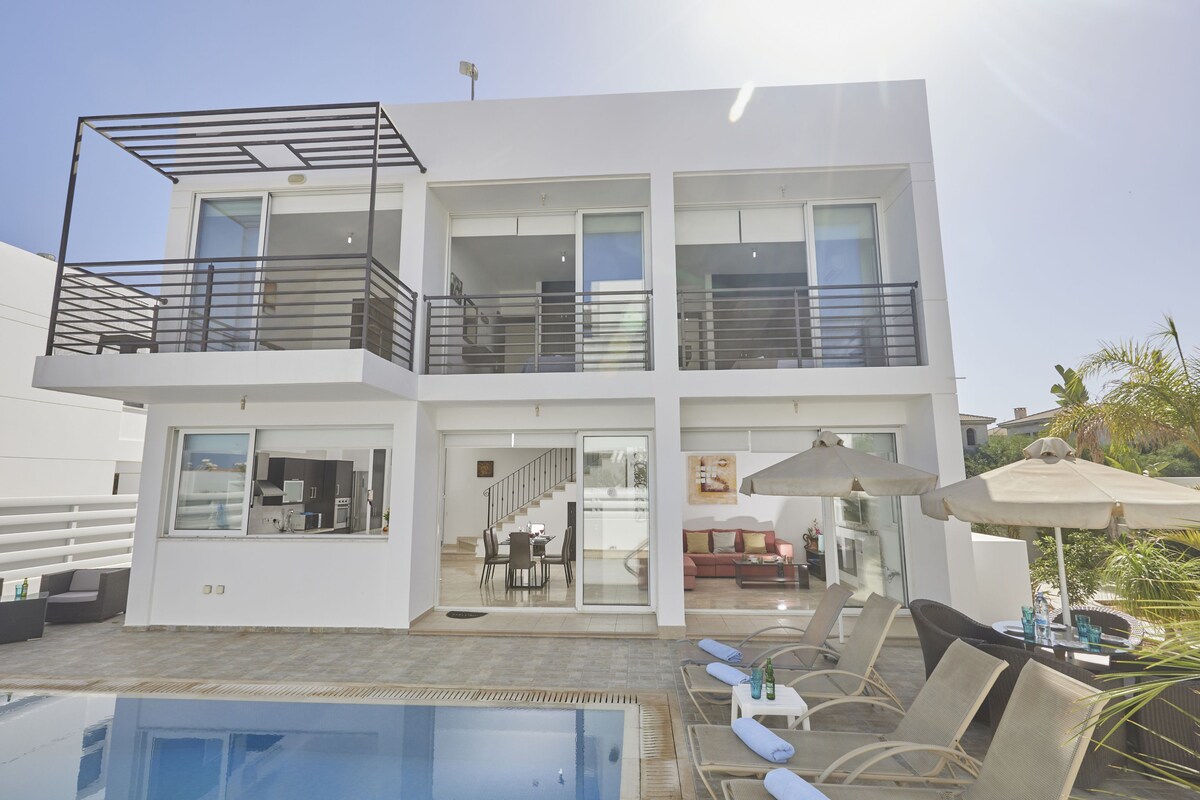 The exterior of the villa highlights a modern two-story structure with large glass doors, allowing views of the pool and garden area. Sun loungers are positioned beside the pool, while shaded seating is provided by umbrellas, creating a relaxing outdoor environment.