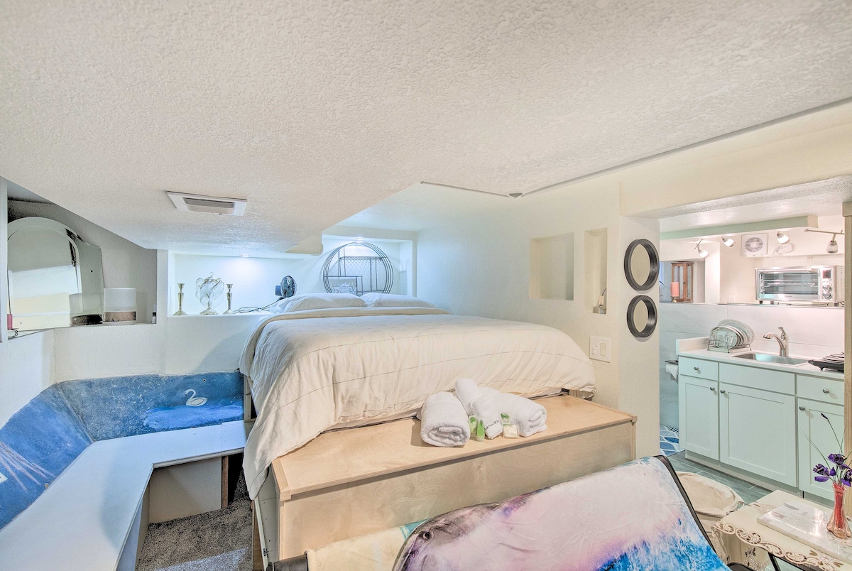 The studio layout features a lofted bed positioned above a cozy sitting area. A kitchenette is visible, equipped with white cabinetry and modern appliances. Soft, ambient lighting enhances the serene atmosphere, while plush towels are neatly arranged on the bed.