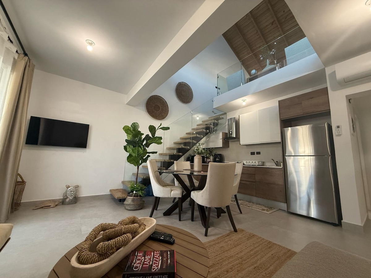 A modern living space features a dining table with four upholstered chairs, accompanied by decorative plants. A staircase leads to an upper level, while light-colored cabinetry and stainless steel appliances are visible in the kitchen area. Natural light filters through the large windows.