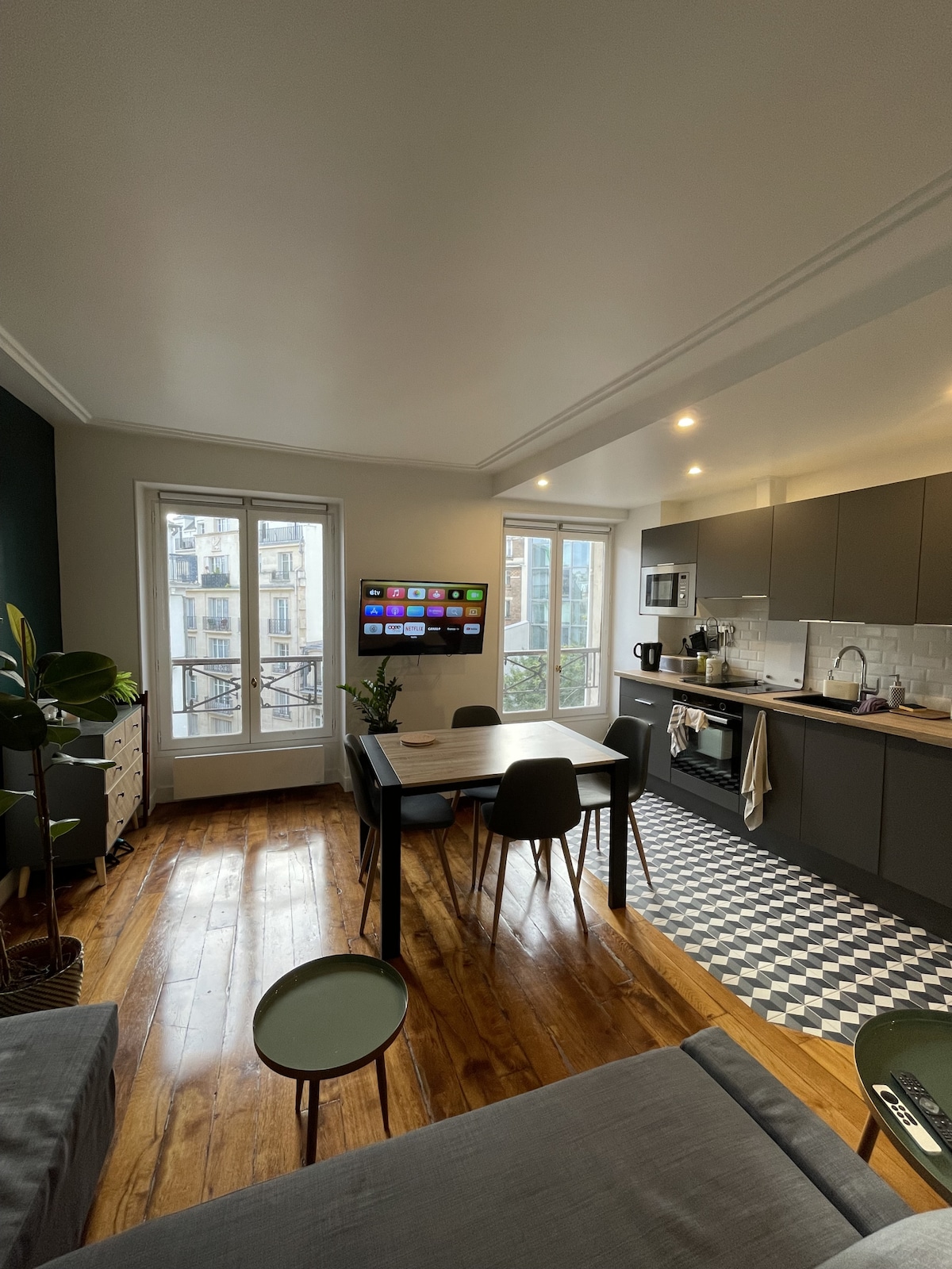 A modern kitchen and dining area is presented, featuring a sleek gray design with a large window allowing natural light. A compact dining table with four chairs is situated beside a mounted television. The tiled floor adds a touch of character to the light-filled space.