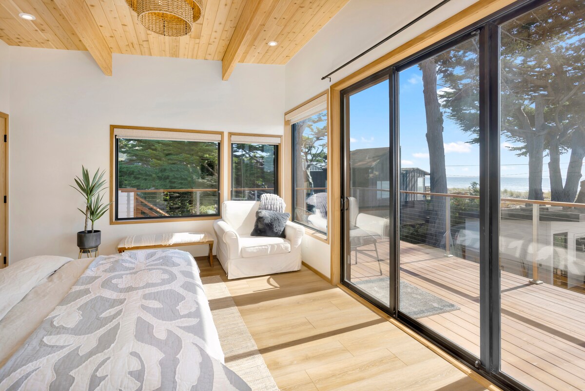 A bright bedroom features large windows offering ocean views and natural light. The space includes a spacious bed adorned with a patterned coverlet, a cozy armchair, and warm wooden ceiling beams, complementing the inviting atmosphere of the room.