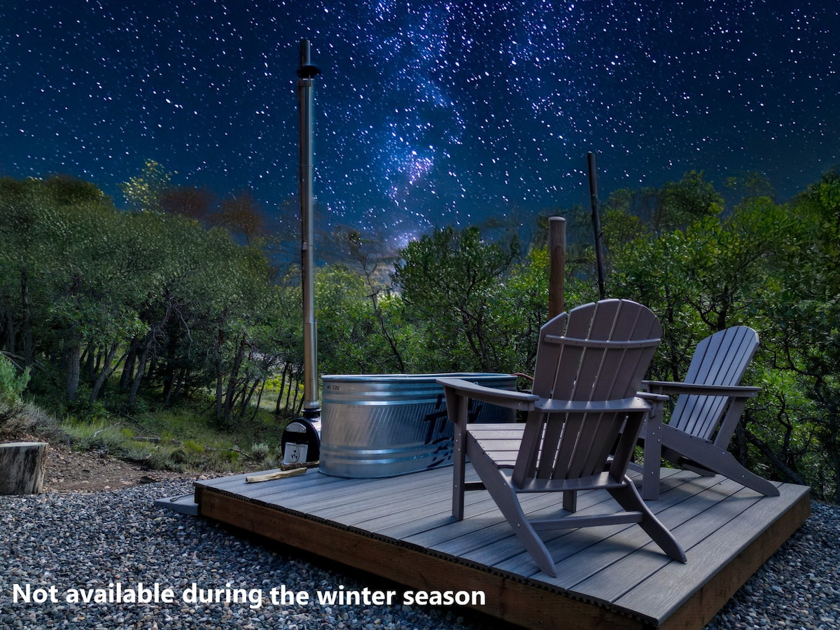 A wooden deck features two Adirondack chairs facing a woodfire hot tub, surrounded by greenery under a star-filled night sky. The glimmering stars create a serene ambiance, inviting relaxation in nature's tranquility.