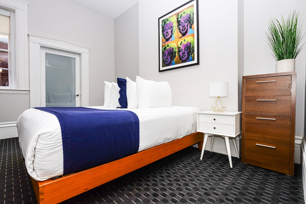 A well-appointed bedroom features a spacious bed with a blue accent blanket. A bedside table with a lamp is positioned next to the bed. A colorful piece of artwork is displayed above the bed, and a tall dresser stands nearby, complemented by a potted plant.