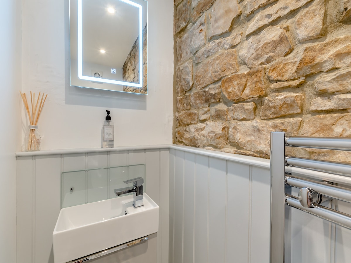 A modern bathroom features a sleek wall-mounted sink and a heated towel rail. The space is illuminated by soft overhead lights, highlighting the textured stone wall and light-colored paneling. A backlit mirror above the sink offers additional brightness and a contemporary touch.