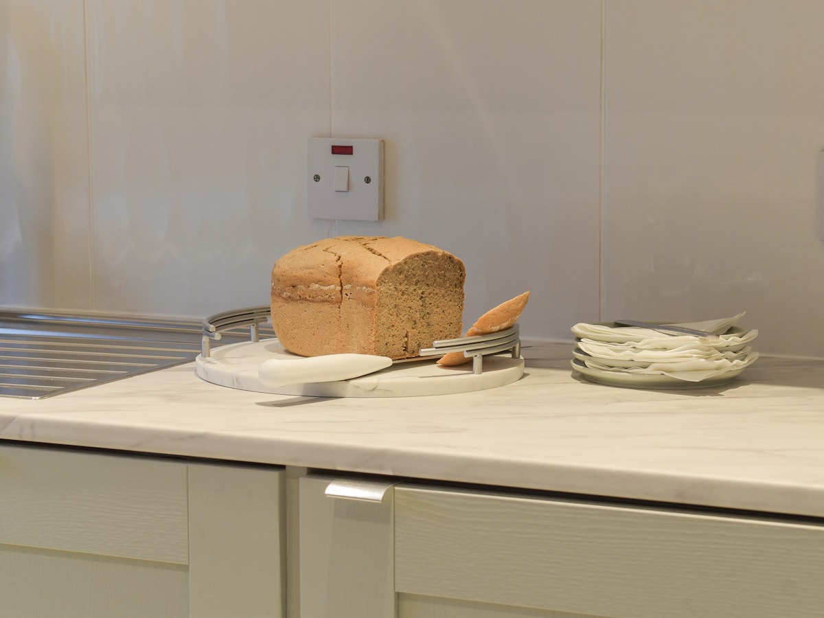 A loaf of bread sits on a round white tray, accompanied by a small wooden knife and a stack of white plates. The kitchen countertop is light-colored and smooth, with a contrasting backsplash partially visible.