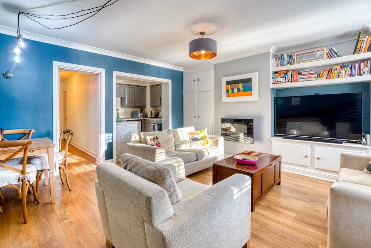 A spacious living area is featured, with light hardwood flooring and blue accent walls. Two comfortable sofas and a coffee table are arranged around a large television. A dining table with wooden chairs is visible, and shelves filled with books add character to the room.
