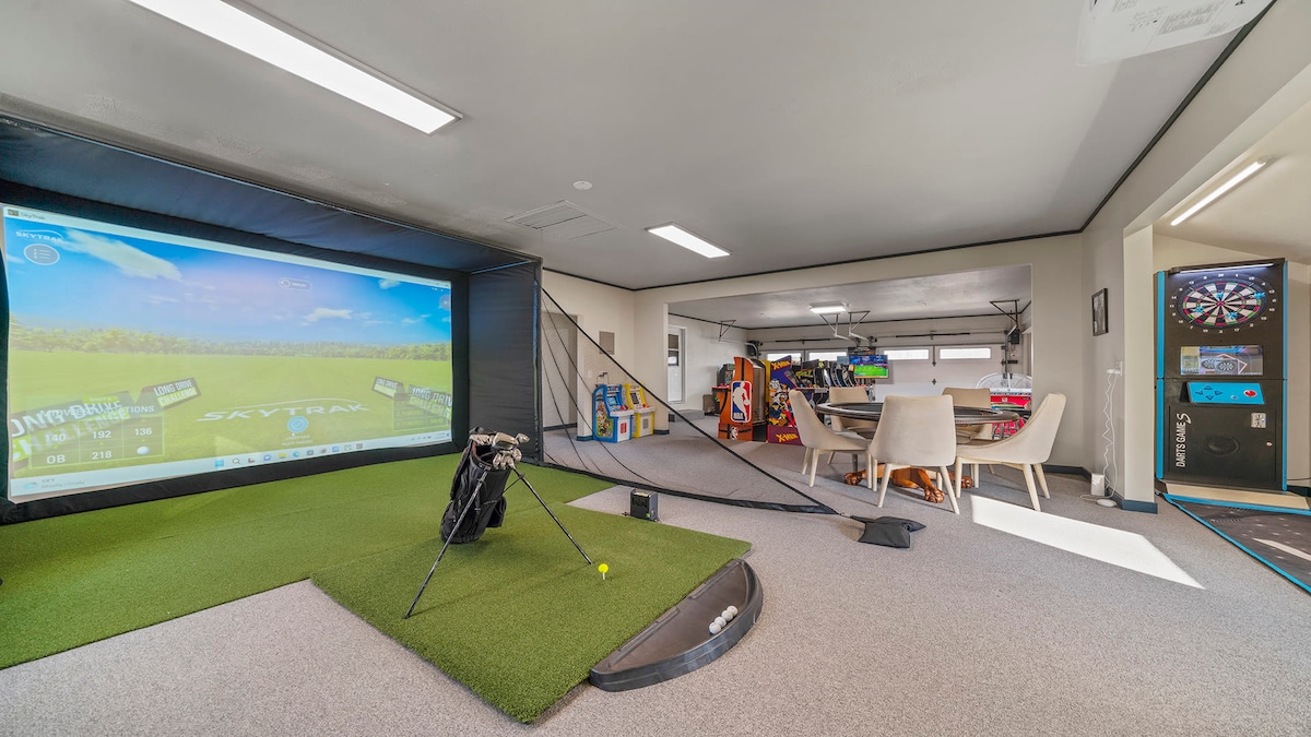 A spacious entertainment room features a golf simulator with a green turf area and a golf bag with clubs beside it. To the right, a 90s arcade gaming area is visible, showcasing colorful arcade machines and a dartboard, creating an inviting atmosphere for fun.