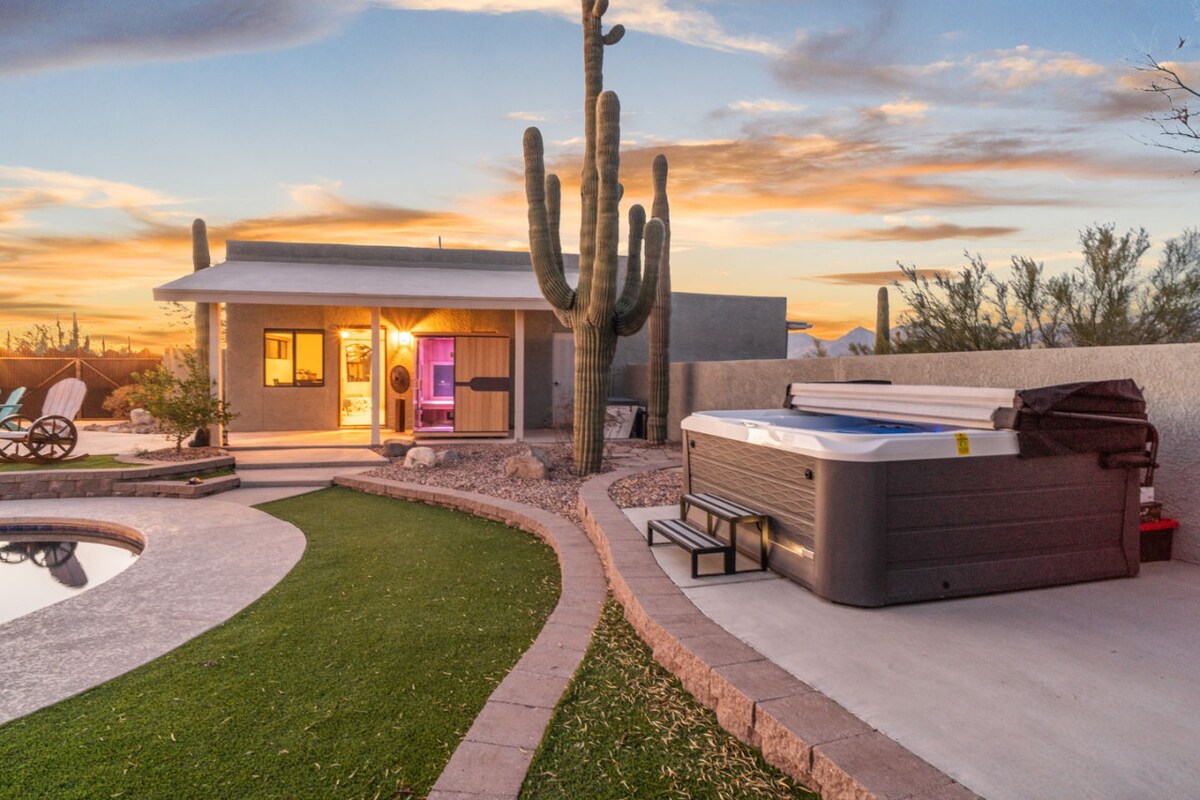 A serene outdoor space is presented with a spa and a swimming pool surrounded by landscaped pathways. A prominent cactus stands beside the property, and the sky displays a gradient of warm colors at sunset, enhancing the tranquil setting.