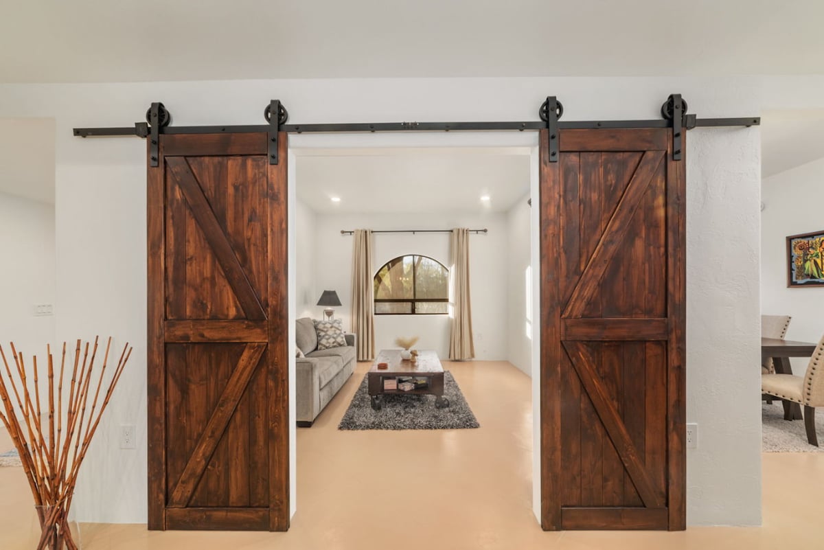 A warm and inviting entrance features large wooden barn doors that slide open to reveal a spacious living area. Natural light enters through an arched window, illuminating the soft seating and stylish decor. Decorative elements such as a floor vase are strategically placed for added charm.