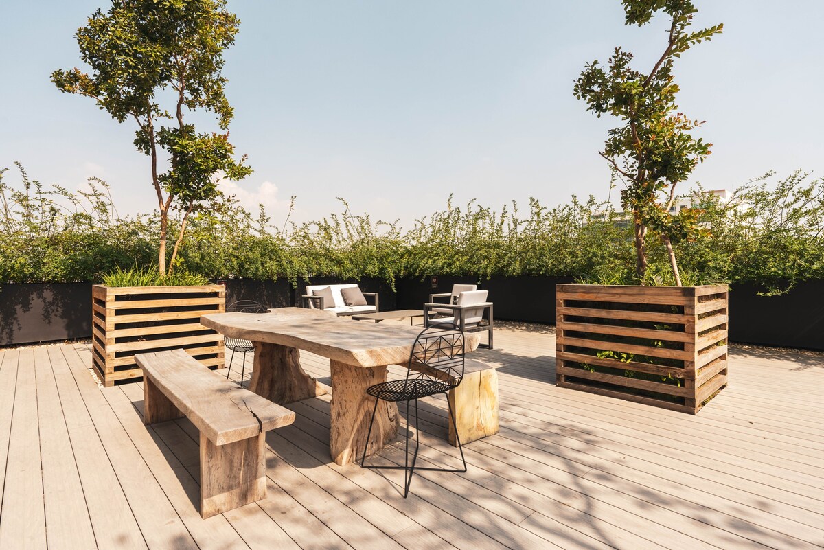 An outdoor terrace is presented with wooden seating arrangements, including a rustic table surrounded by various chairs. Green plants are contained in modern planters, providing a natural backdrop against the clear sky, creating a tranquil space for relaxation.