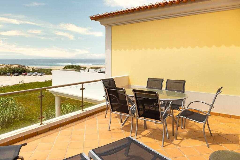 A private terrace is showcased with a dining table surrounded by six chairs, inviting outdoor meals. The area features a clear view of the beach and sea in the background, complemented by a smooth tiled floor and partial glass railing for safety.