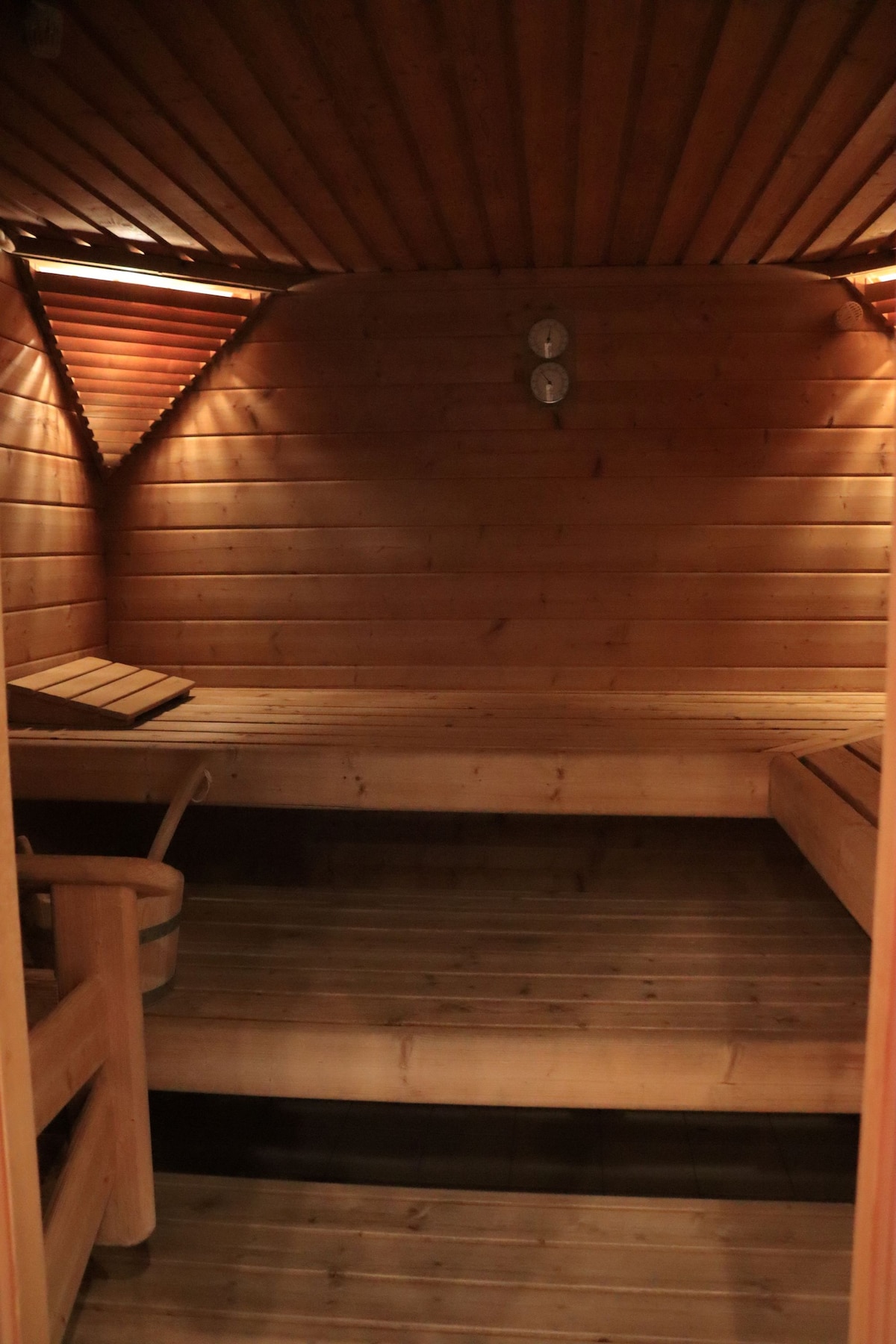 A wooden sauna interior is shown, featuring cozy bench seating arranged against the walls. Soft lighting highlights the warm wooden tones, creating a calming environment. Two temperature gauges are mounted on the wall, and the floor consists of slatted wooden boards.