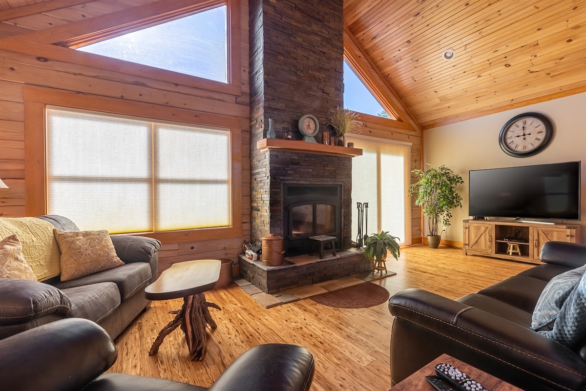 The living room features a vaulted wooden ceiling with tall windows allowing natural light to fill the space. A stone fireplace serves as the focal point, while comfortable seating is arranged around a wooden coffee table. A television sits across from the seating area.