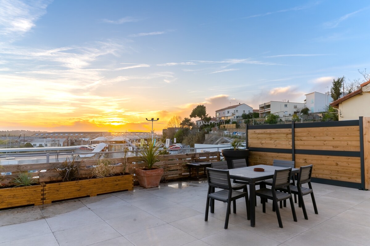 A spacious rooftop terrace features a dining table surrounded by black chairs, set against a backdrop of a vibrant sunset. Potted plants are positioned along the perimeter, and the expansive sky displays hues of orange and blue, creating a serene outdoor space.