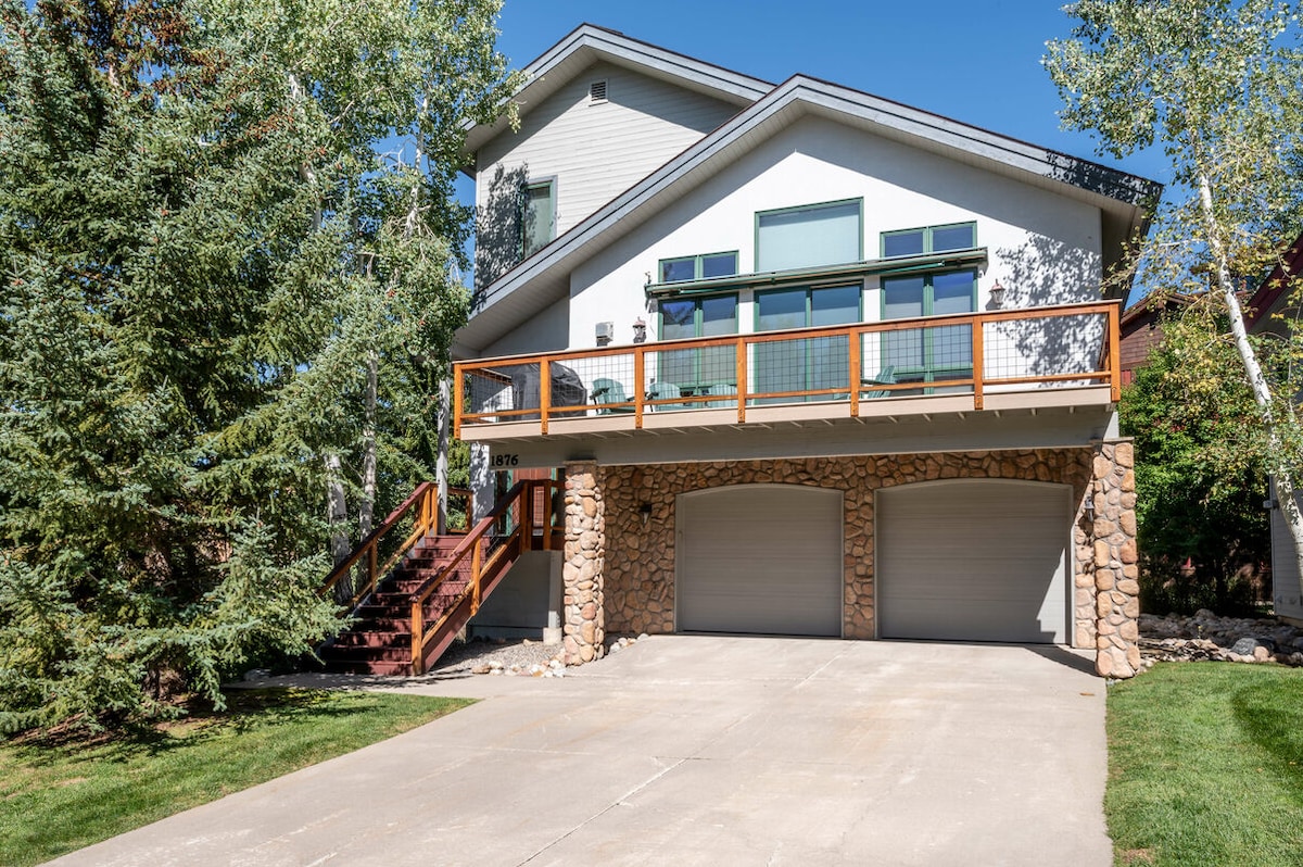 A modern two-story home features a charming wood and stone exterior with large windows, allowing natural light to fill the space. A spacious deck provides outdoor seating and access to the front entrance. A paved driveway offers parking for up to four vehicles.