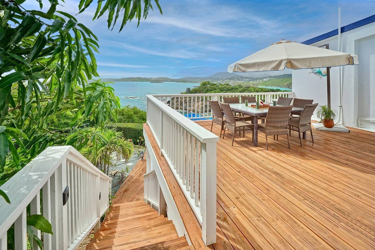 A wooden deck with a dining table and chairs is positioned under a large umbrella, offering views of the Caribbean Sea. Lush greenery is visible along the edges of the deck, which is accessed by steps leading down. The sunlit scene highlights the tranquil outdoor setting.