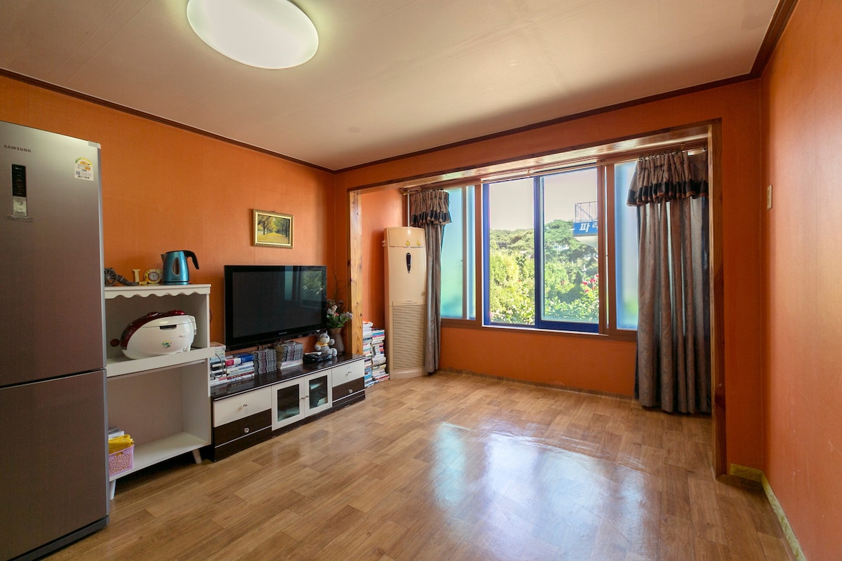 A bright living area features orange walls and large windows providing views of the outside greenery. A television and refrigerator are visible alongside a white cabinet, while light hardwood flooring enhances the spacious feel of the room.