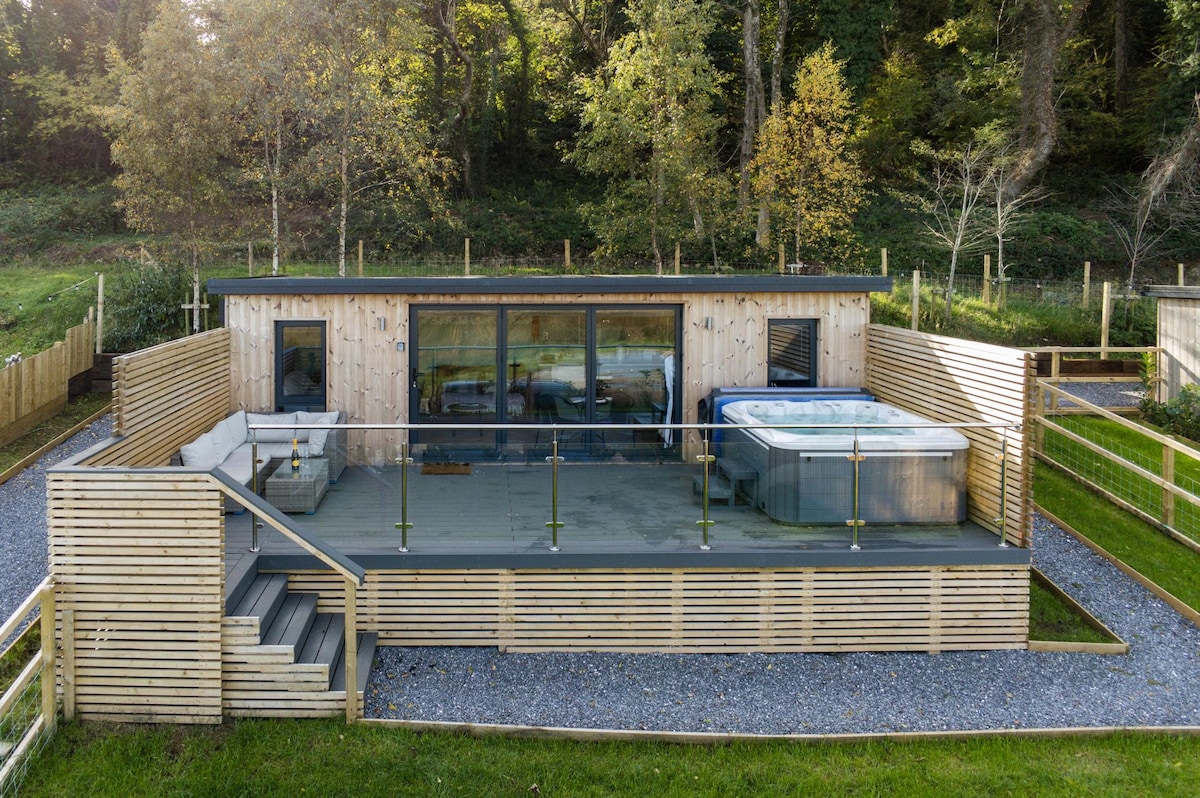 A modern cabin is depicted with a spacious wooden deck and a private hot tub. Expansive glass doors lead into the interior, providing a view of the surrounding greenery. A railing encloses the deck, enhancing the tranquil outdoor space.