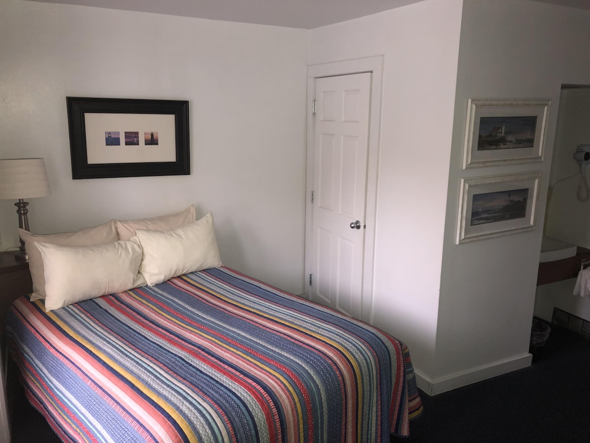 A cozy room features a full-size bed with a colorful striped blanket. A nightstand with a lamp is positioned beside the bed. Framed artwork adorns the wall above the bed, and a door leads to the ensuite area visible in the background.
