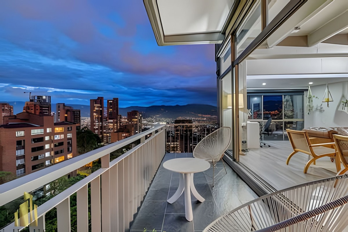A private balcony offers sweeping views of the city skyline and surrounding mountains. Modern outdoor furniture, including a round table, is displayed on the tiled floor. The glass doors provide a seamless connection to the interior space, illuminated and inviting in the evening light.