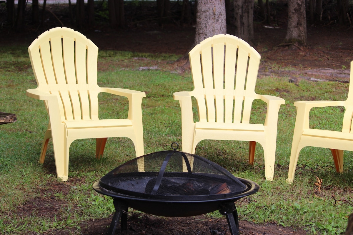 A fire pit surrounded by four yellow Adirondack chairs is depicted on a grassy area. The fire pit features a metal grating on top, with a heartening setup inviting a gathering. Trees provide a backdrop, contributing to a natural setting.