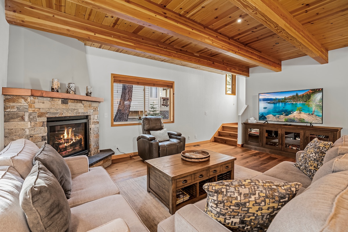 The living room features a stone fireplace, offering a cozy focal point. Comfortable seating is arranged around a wooden coffee table, with a television mounted on the wall. Large windows provide natural light, complementing the rustic wooden beams overhead.
