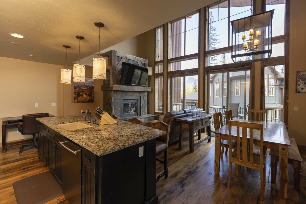 A spacious kitchen area features a dark granite island with modern appliances and a set of kitchen knives. Natural light floods in through large windows, highlighting a dining table and chairs nearby. A cozy fireplace offers warmth to the inviting space.