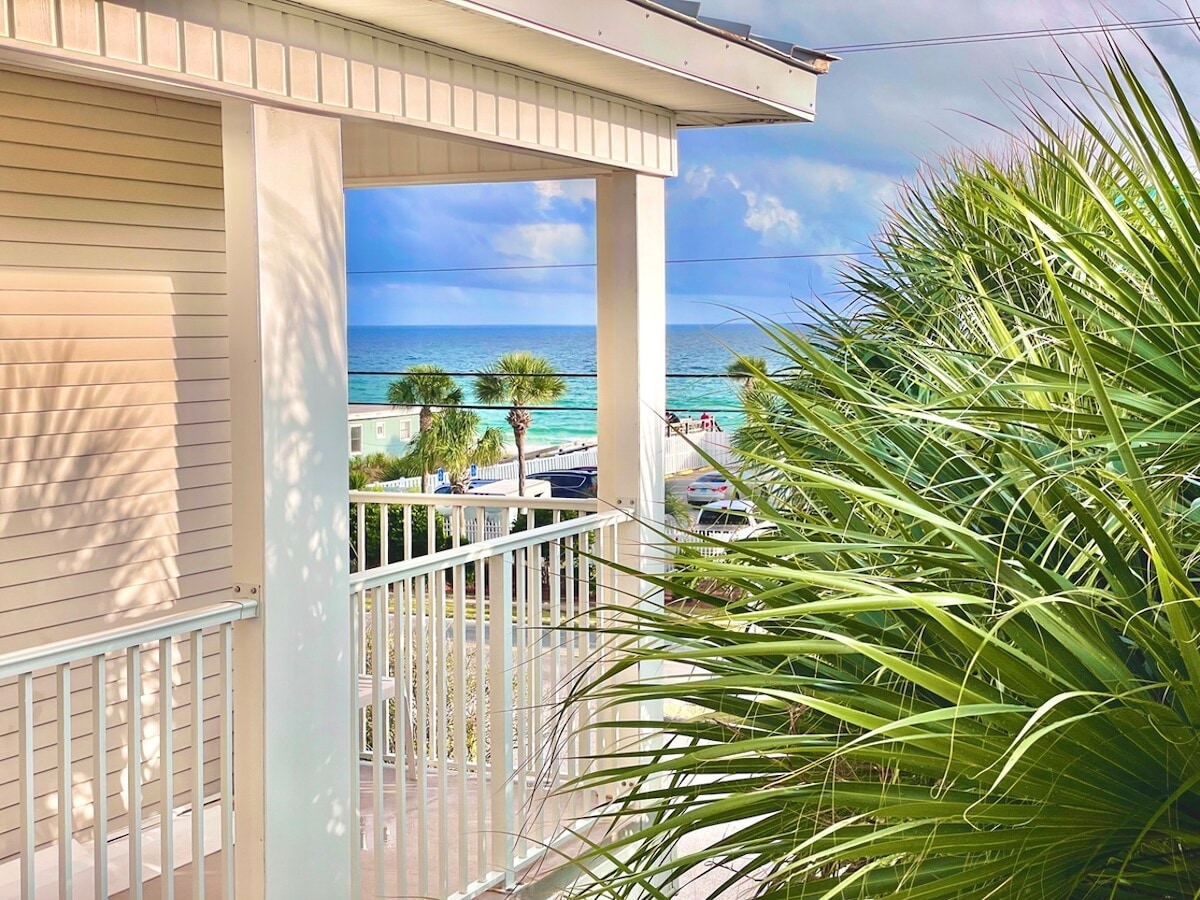 A serene view from a private balcony is framed by lush palm leaves, overlooking the ocean waters. The beach is visible in the distance, with soft waves and a peaceful coastline. The sky is partly cloudy, adding to the tranquil ambiance.