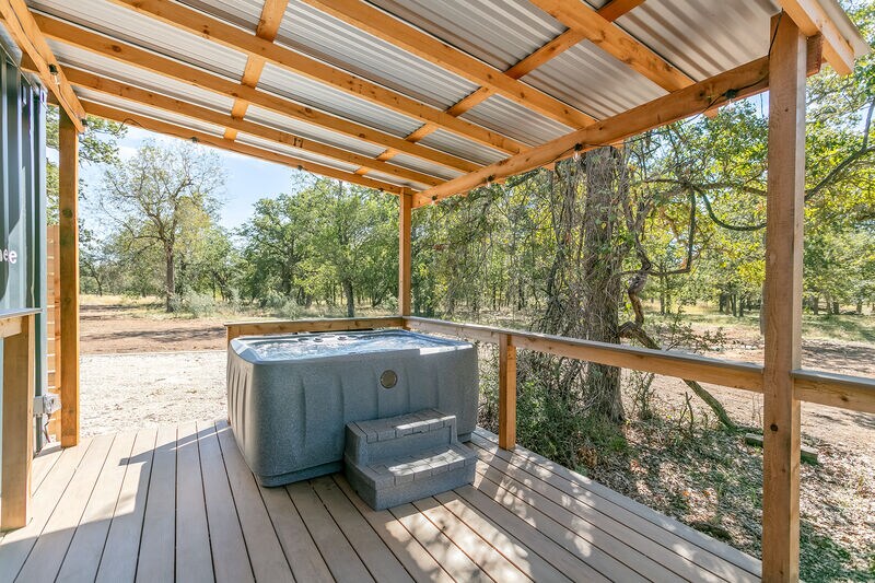 An outdoor deck features a private hot tub, surrounded by a wooden railing and steps for easy access. Natural sunlight highlights the spacious area, set against a backdrop of trees and open space, offering a tranquil environment for relaxation.